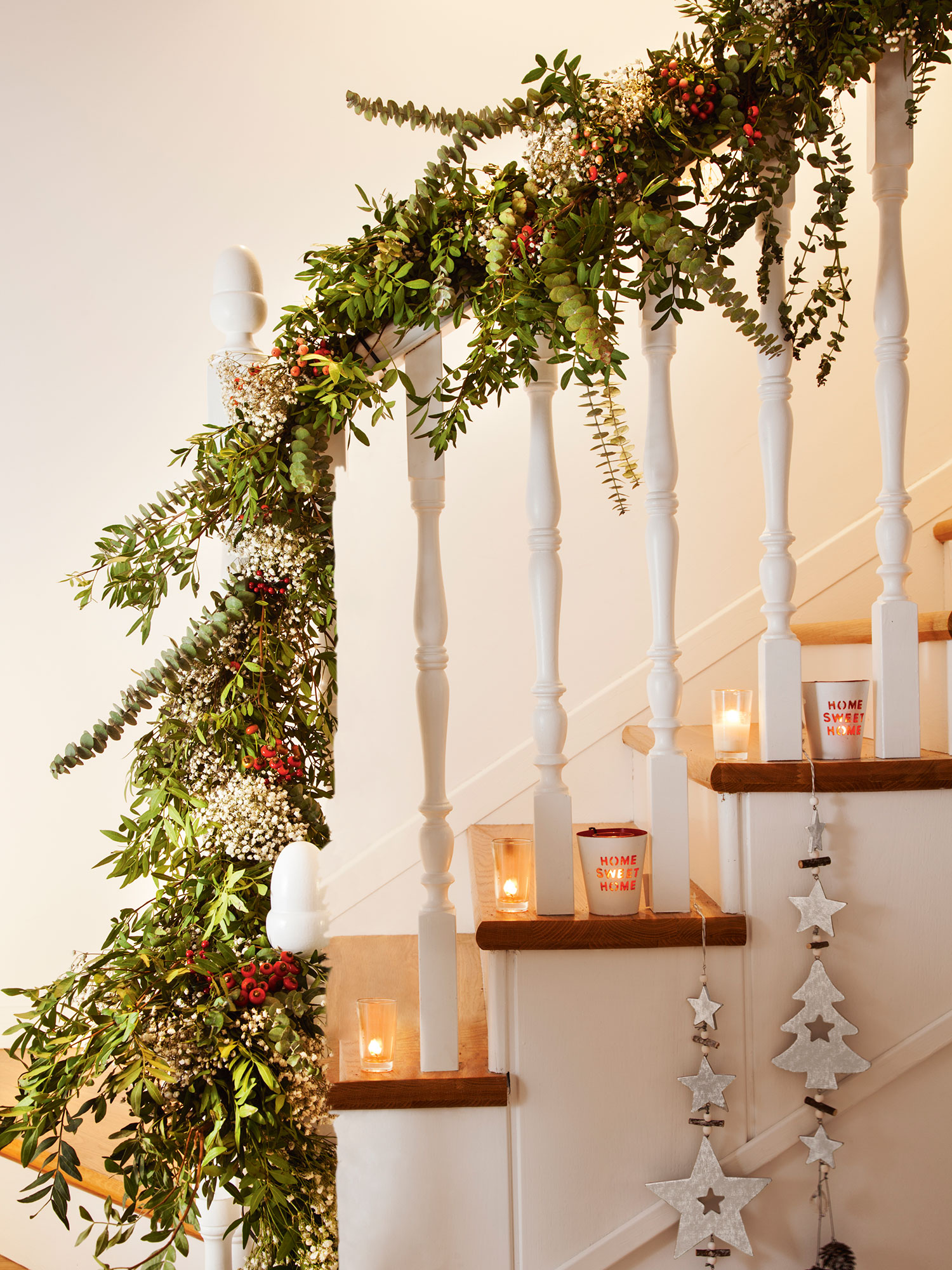 Detalle de escalera con peldaños de madera y barrotes blancos decorada de Navidad con ramas verdes, adornos y velas