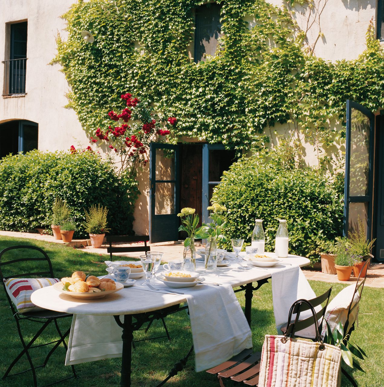 Comedor exterior frente a la casa con una gran mesa en tonos blancos con patas de hierro. 