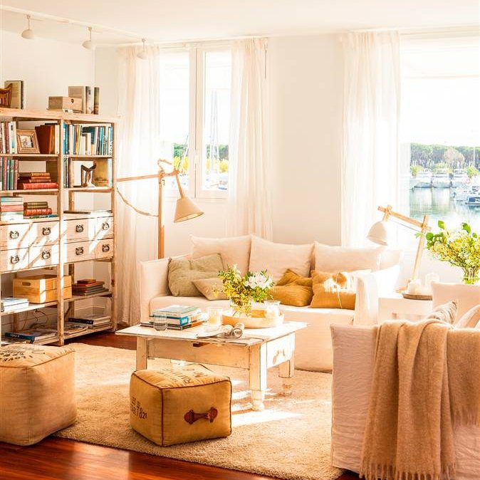 Salón con sofás blancos, alfombra, puffs vintage, lámparas Tolomeo, estantería y finas cortinas blancas