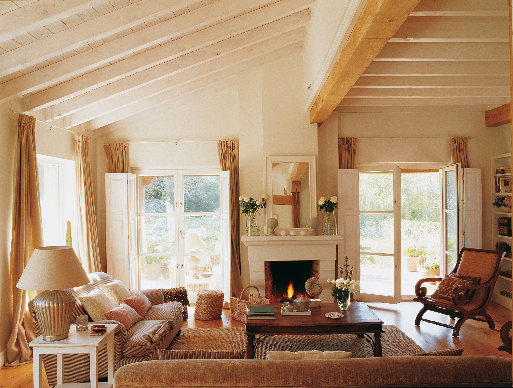 Salón de casa de campo en blanco-beige con vigas y chimenea y ventanales al exterior.