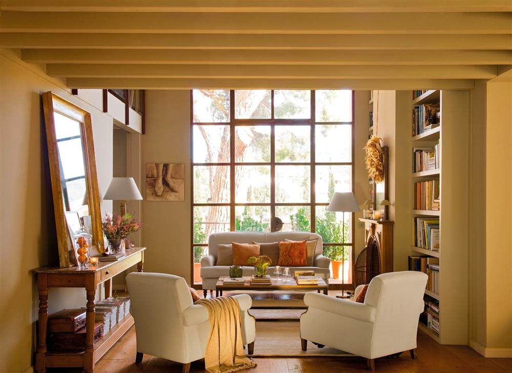 Salón ejemplo de distribución con sofá y butacas enfrentados con mesa de centro, consola con espejo, paredes en arena y librerías de obra junto a gran ventanal