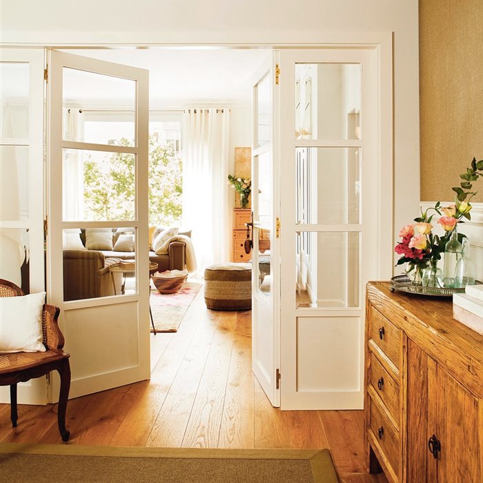 Recibidor conectado al salón con puerta de doble hoja con cuarterones de cristal en blanco, parquet de madera, cómoda de madera envejecida con tiradores metálicos