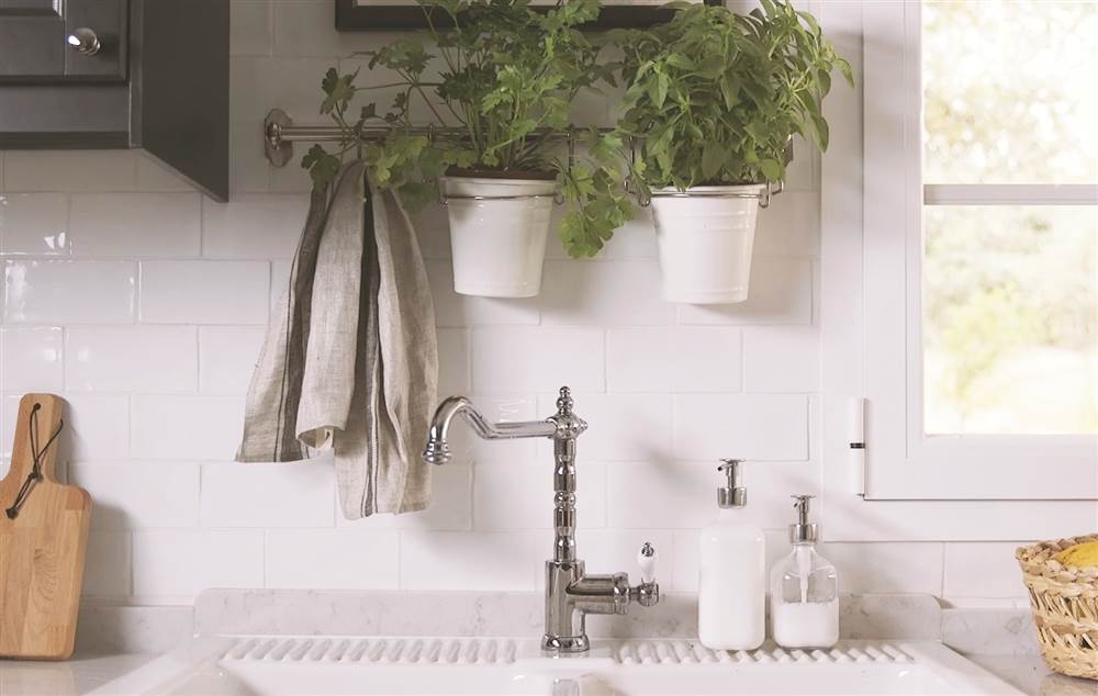 Zona de aguas de la cocina con antepecho de azulejos blancos, grifería vintage, riel plantas aromáticas colgando, jabones y paño de cocina topo