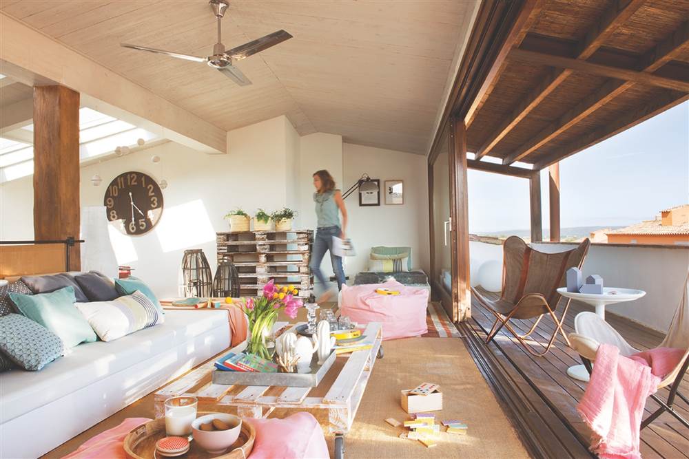 Salón junto a la terraza con sofá blanco, mesa de centro de palé, sillas Butterfly, techo abhuardillado y ventanal corredero