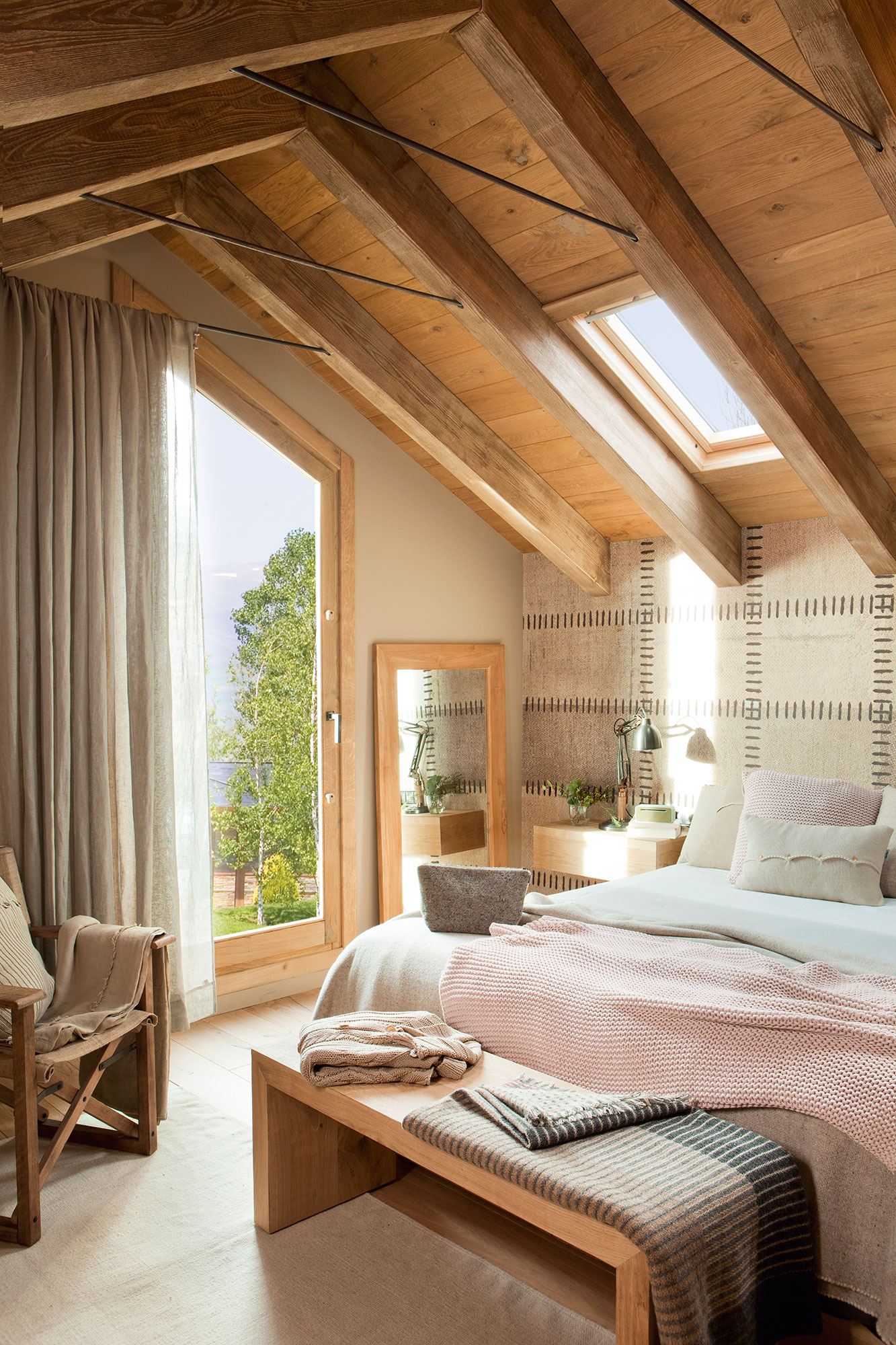 Dormitorio abuhardillado con vigas de madera y cabecero revestido de papel pintado que simula cuero.