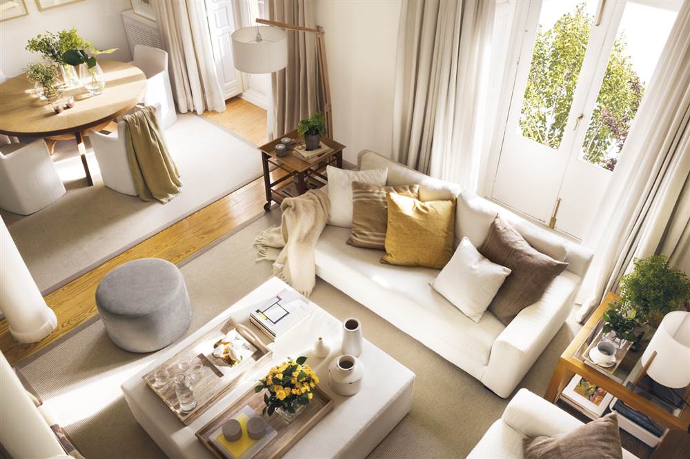 Salón con sofá blanco con cojines marrones, puf de centro cuadrado tapizado en crudo y otro redondo gris