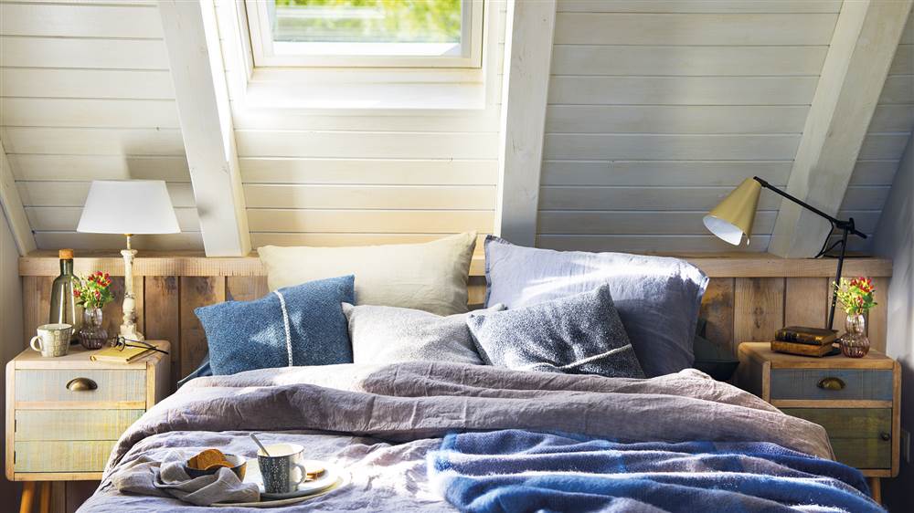 Dormitorio con techo abuhardillado en madera blanca, ventana velux, ropa de cama en tonos azules, mesitas de noche de madera y lámparas de sobremesa