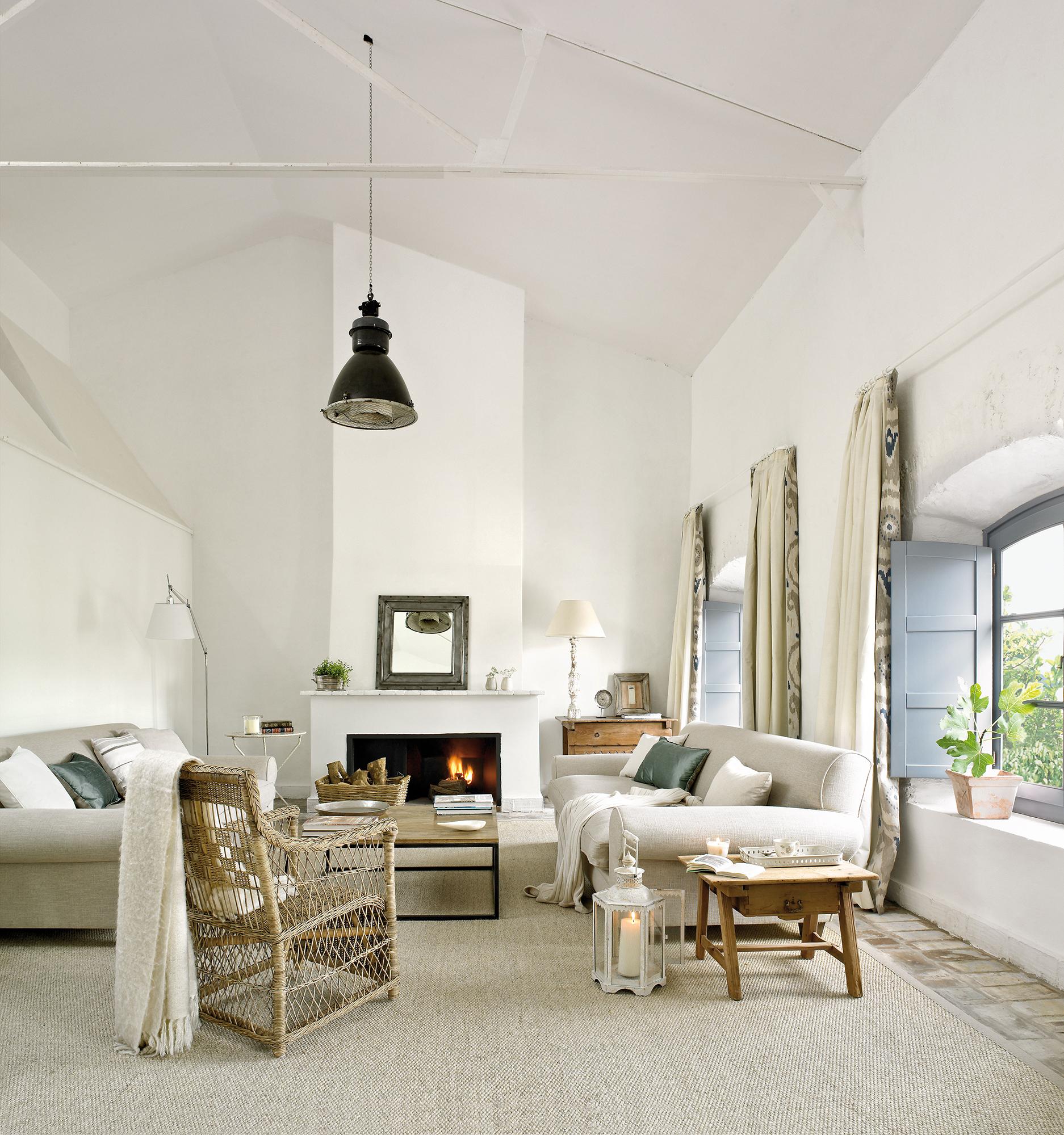 Salón blanco con alto techo abuhardillado, chimenea y butaca de fibra natural.