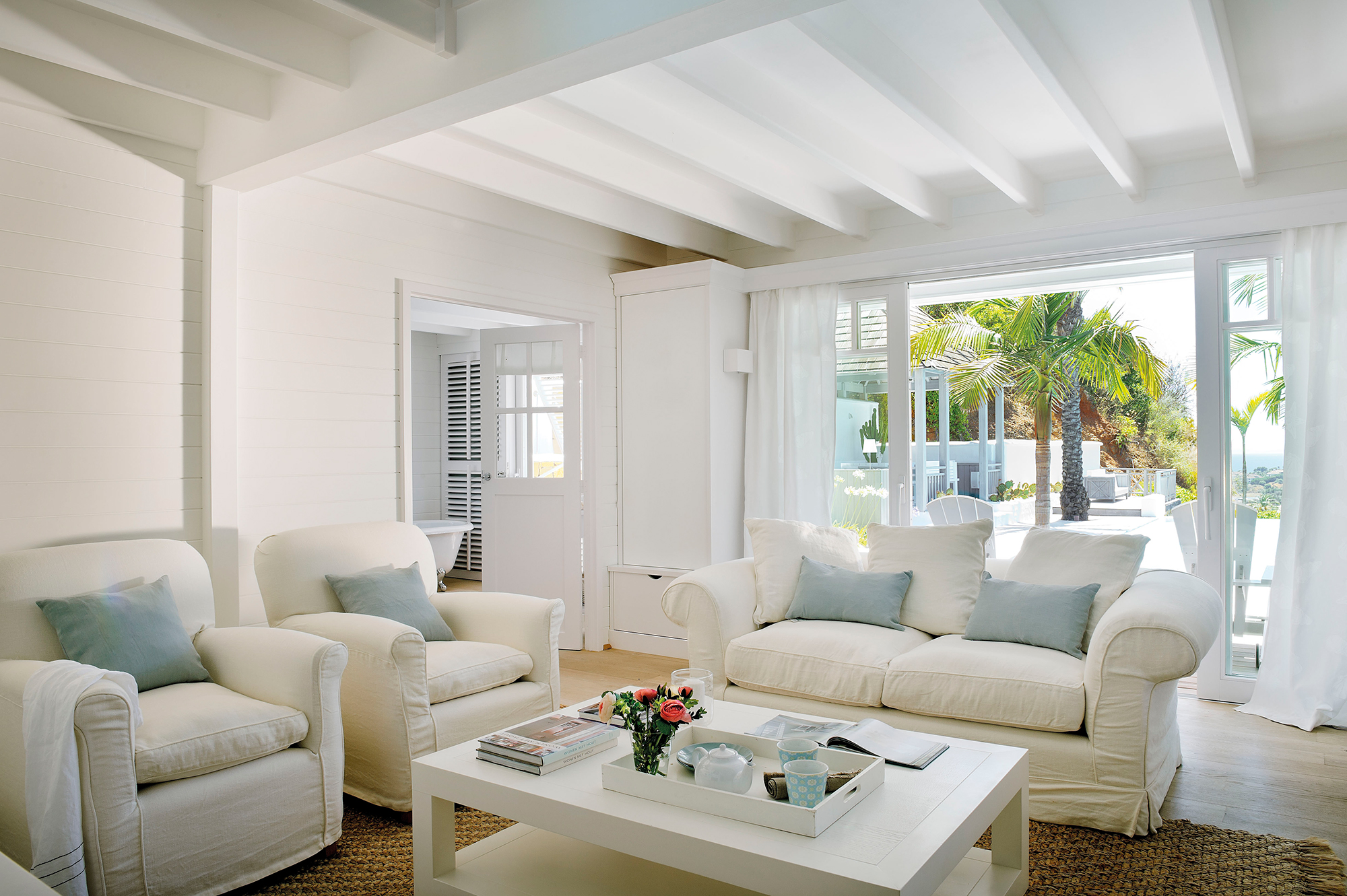 Salón con sofás y butacas blanco, ventanal y mesa de centro blanca.