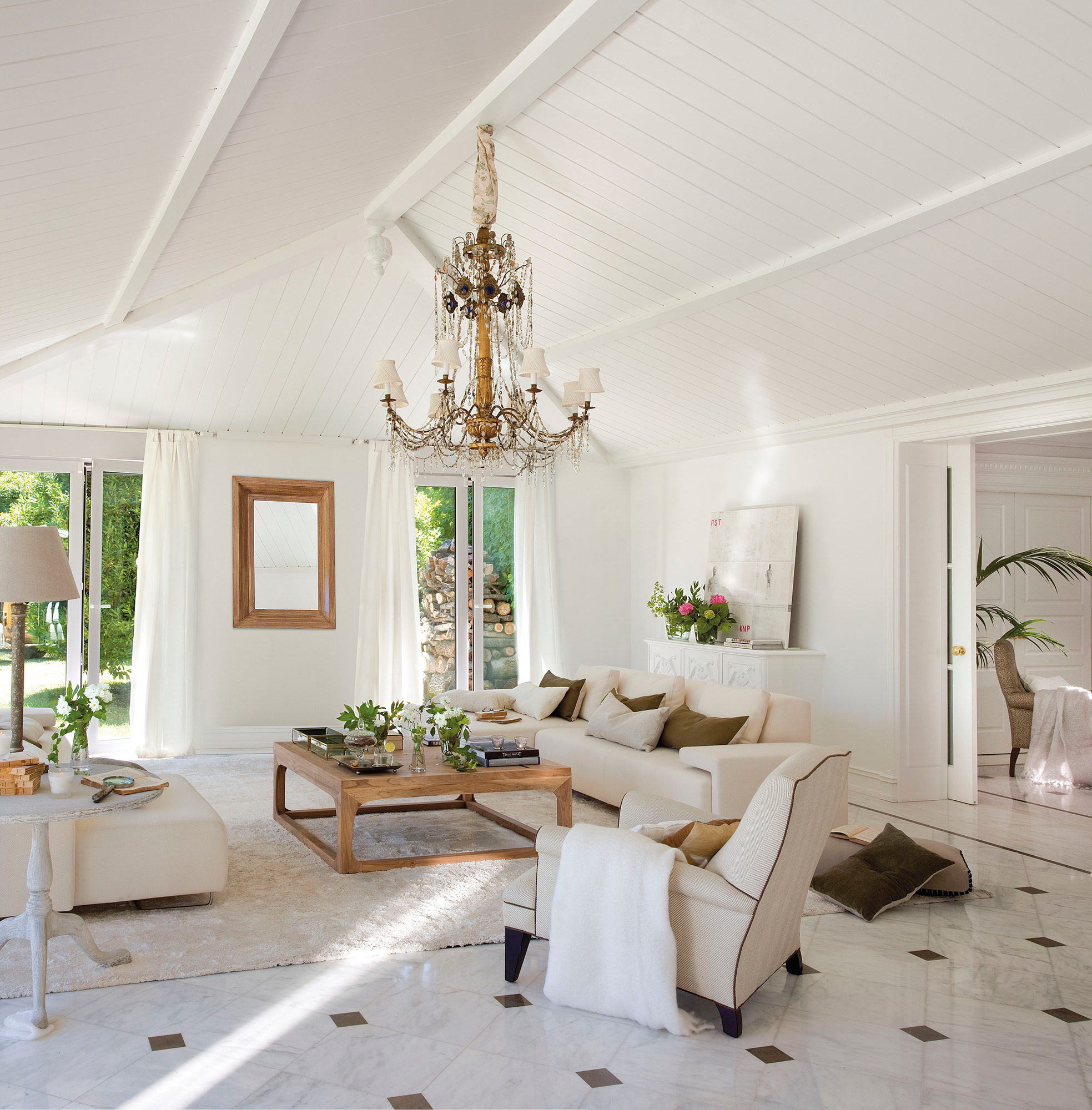 Salón blanco sobre blanco con alfombra, techo abuhardillado, mesa de centro de madera y lámpara de araña.