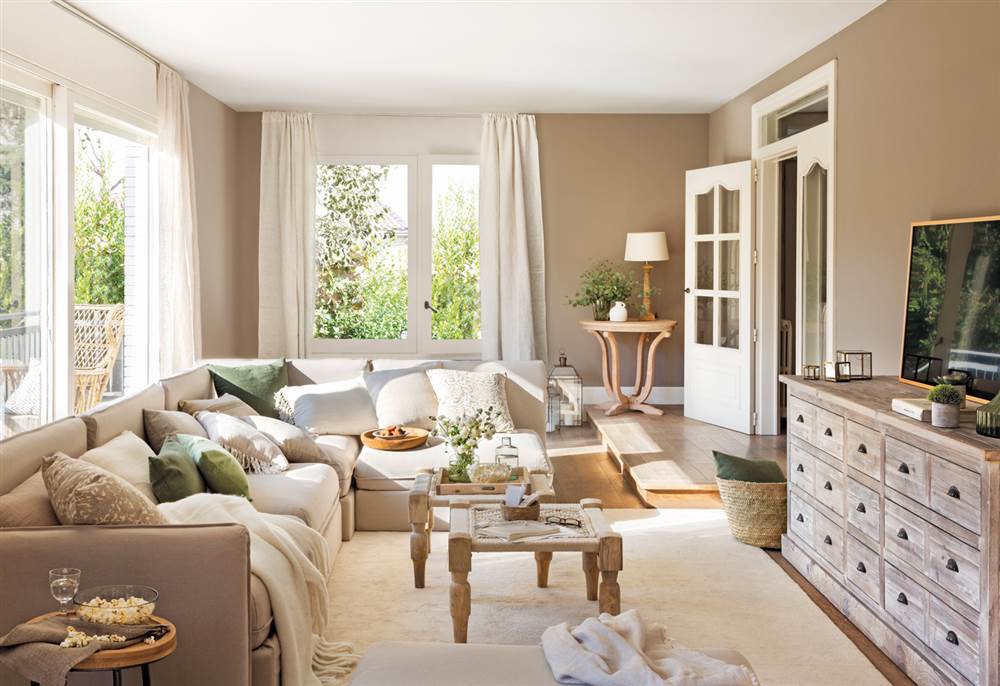 Salón en tonos crema y beige con sofá con chaise longue, aparador de madera, doble mesa de centro y cortinas