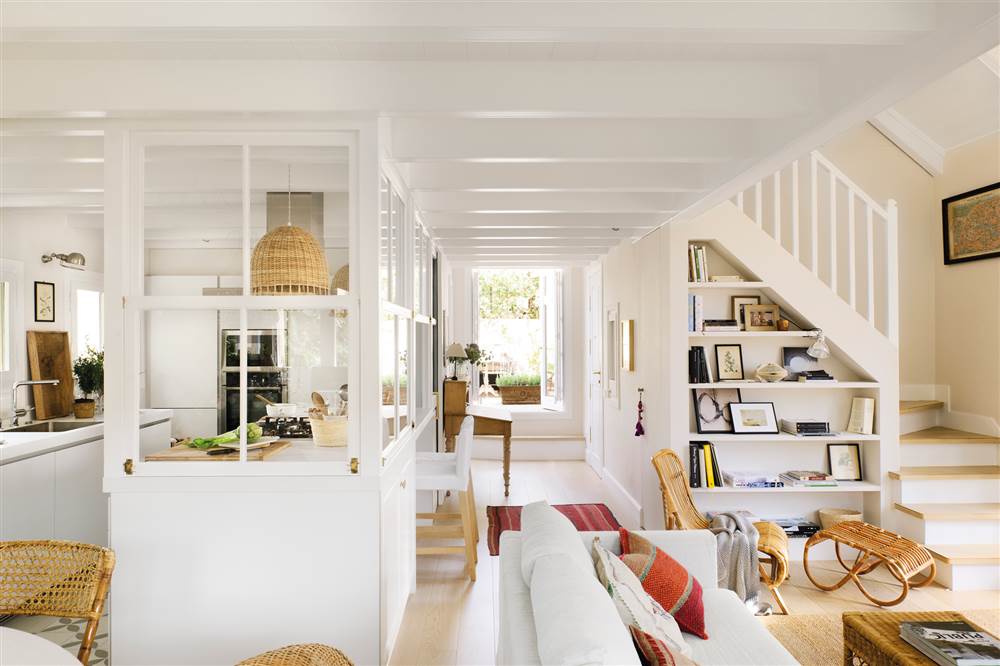 Salón y cocina en blanco con tabique semiacristalado, escaleras con estantería de obra, muebles de caña y fibra natural y techos de madera