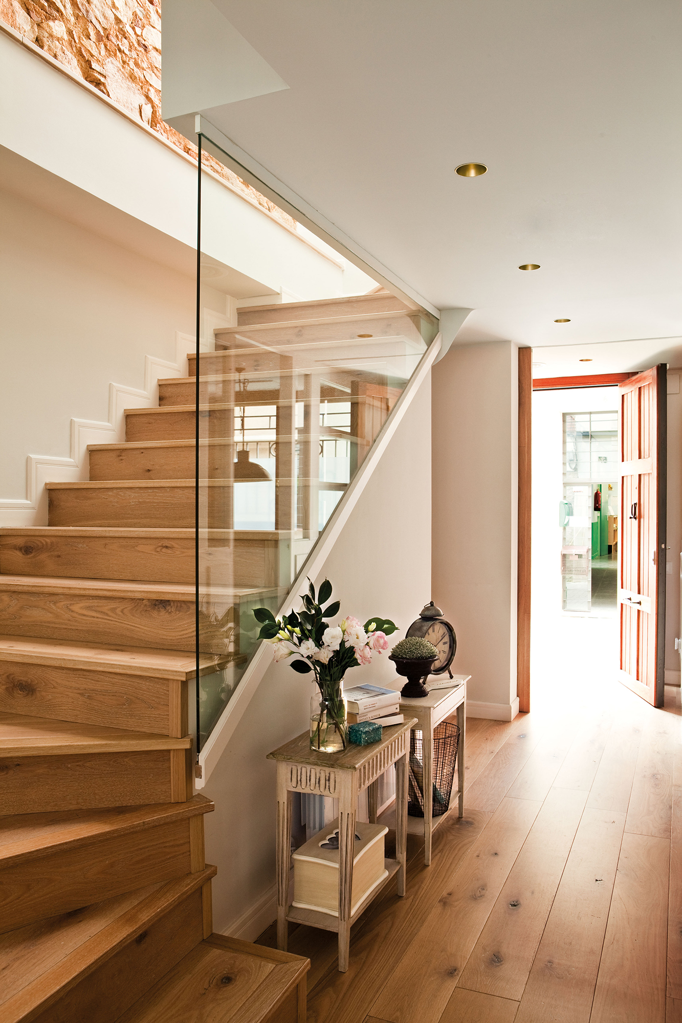 Recibidor con escalera de madera y barandilla de cristal decorado con dos consolas gemelas clásicas