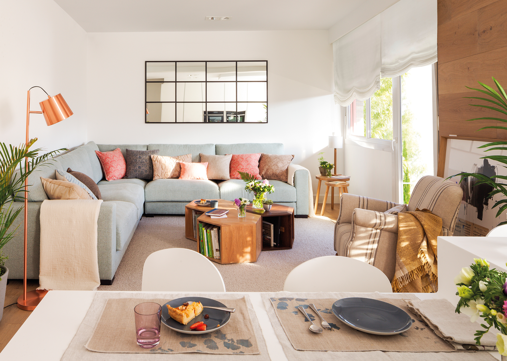 Salón moderno abierto al comedor con sofá esquinero gris, cojines rosas, espejo de cuarterones, mesa de centro de madera y alfombra beige de pelo.