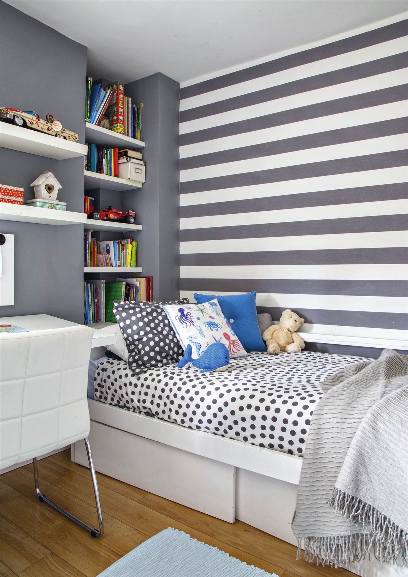 Dormitorio juvenil con papel pintado a rayas, ropa de cama de topos y hueco en la pared con estantes.