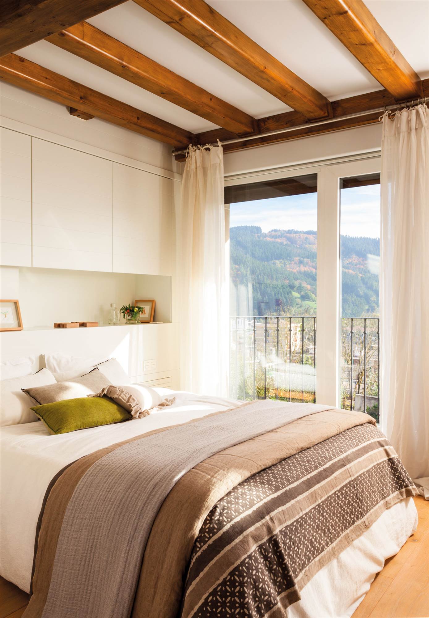 Dormitorio moderno con vigas rústicas y armario a medida en la pared del cabecero. 