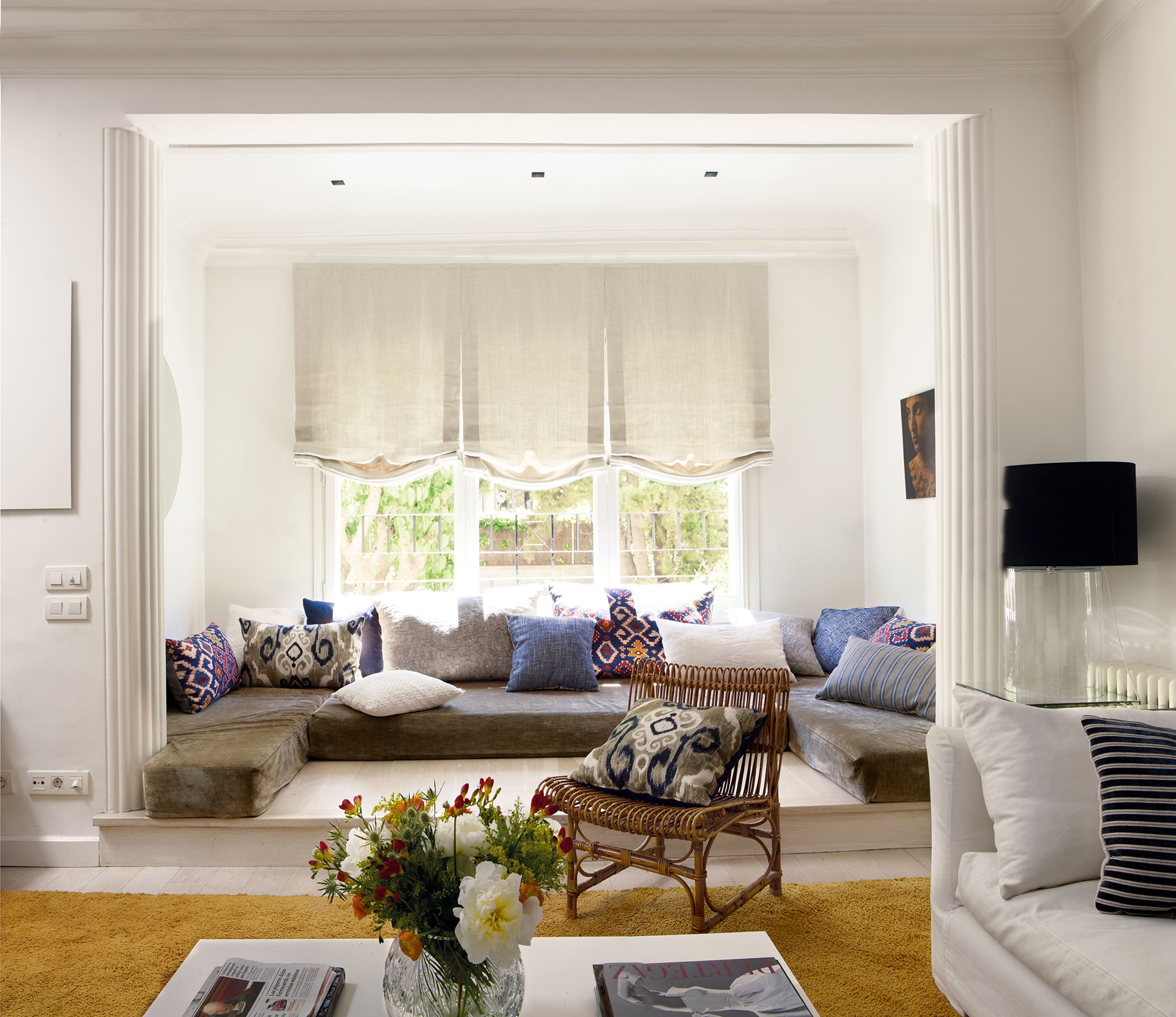 Salón moderno con zona chill out con colchonetas grises en la ventana en forma de L y cojines lilas. 