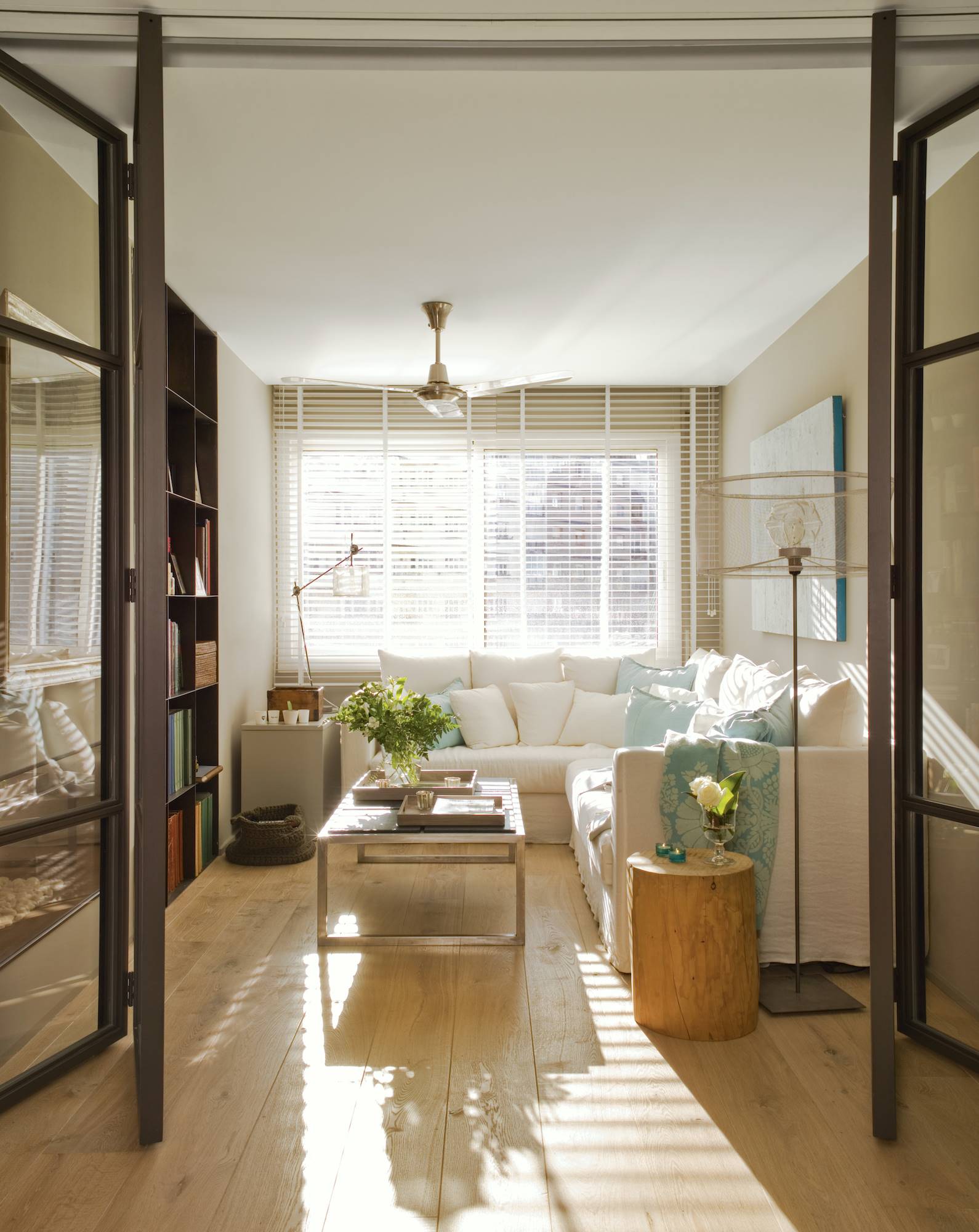Salón con ventana con persiana, sofá, mesita de noche tipo tronco, estantería y puertas de cristal oscuras.