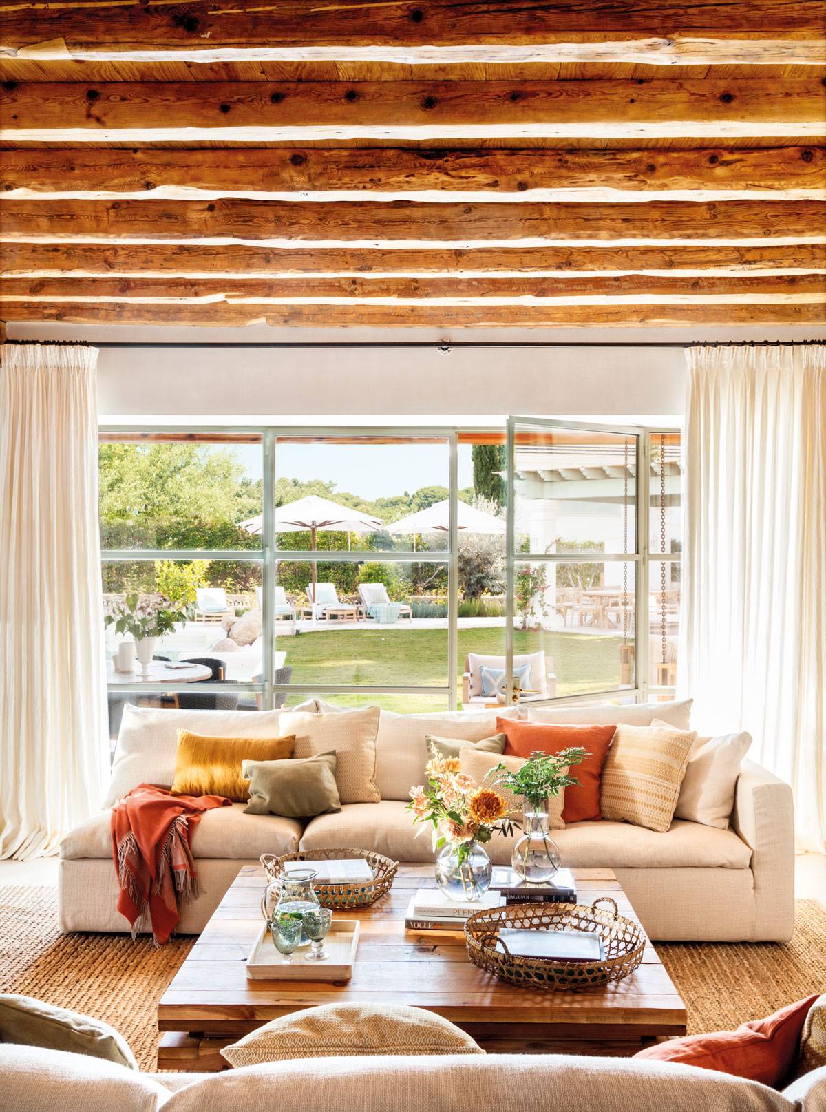 Salón con sofás blancos y vigas de madera y gran pared acristalada con vistas al jardín