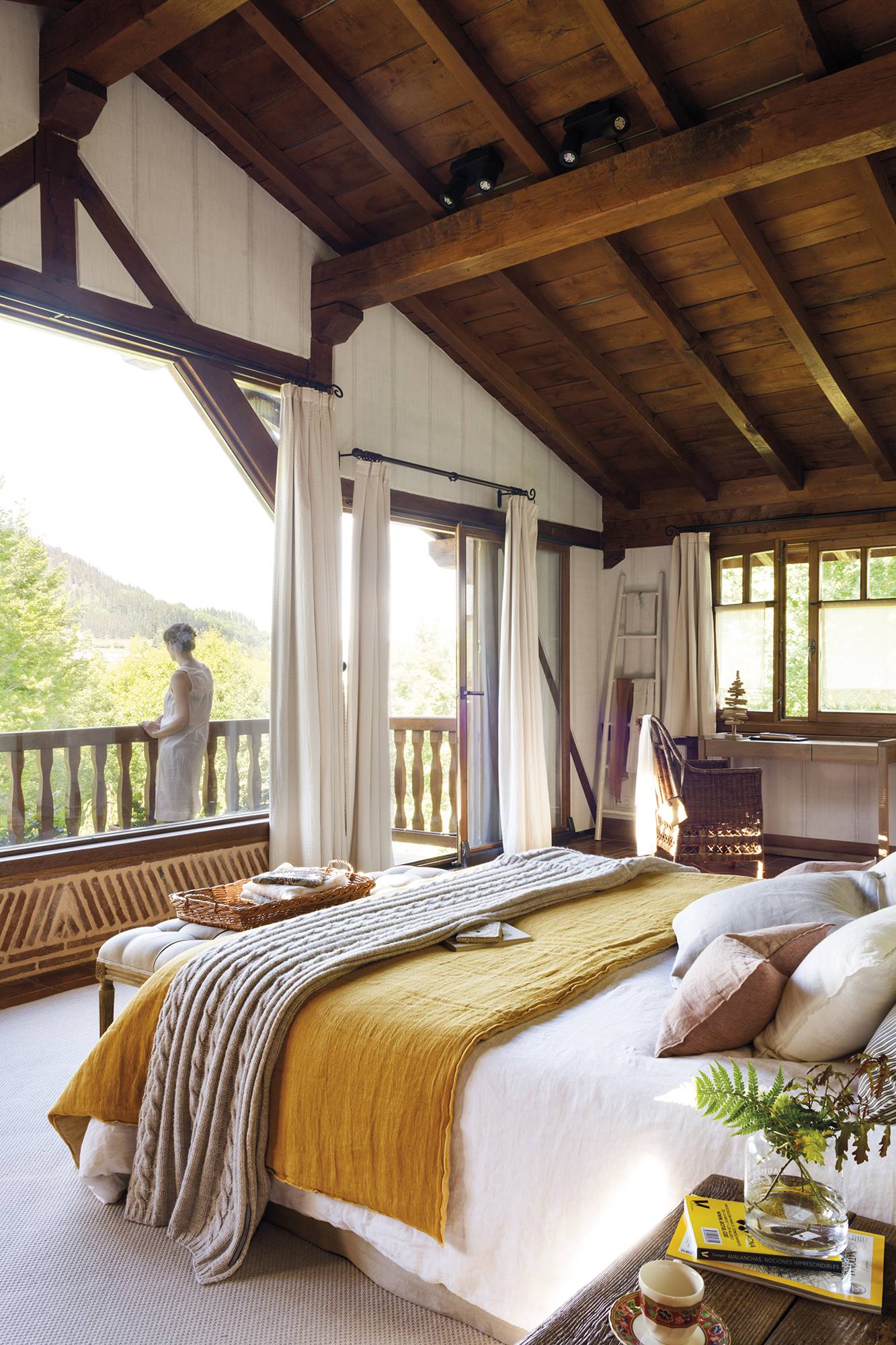 Dormitorio de casa de campo con techo de vigas de madera y gran ventanal con balcón y vistas a las montañas