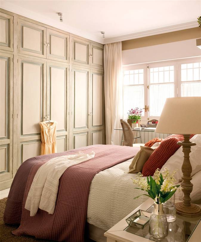 Dormitorio con armario empotrado que ocupa toda la pared con cuarterones, escritorio, cortinas t cama con ropa de cama blanca y granate