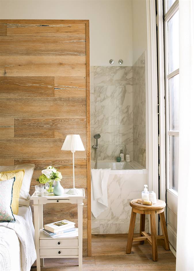 Dormitorio con baño en suite con cabecero de madera, mesita de noche, taburete y vista de la bañera revestida de mármol_395787