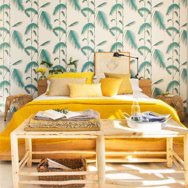 Dormitorio con pared de cabecero empapelada con papel pintado de motivos vegetales, ropa de cama y detalles en amarillo, banqueta de madera, mesitas de noche de fibra natural y ce