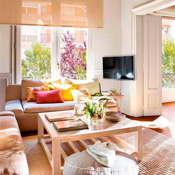 Salón con televisor colgado en la pared, mesa de centro de madera, sofás con cojines de colores y estor enrollable.