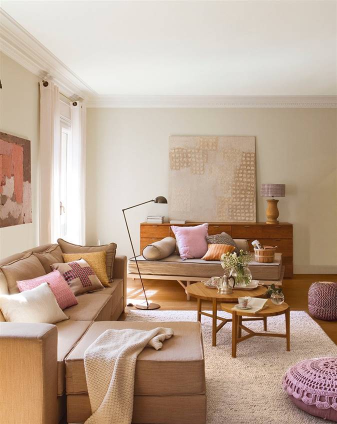 Salón en tonos claros y beige, con sofá marrón, mesas nido de centro redondas, pufs rosas, alfombra, aparador vintage, cuadro y lámparas