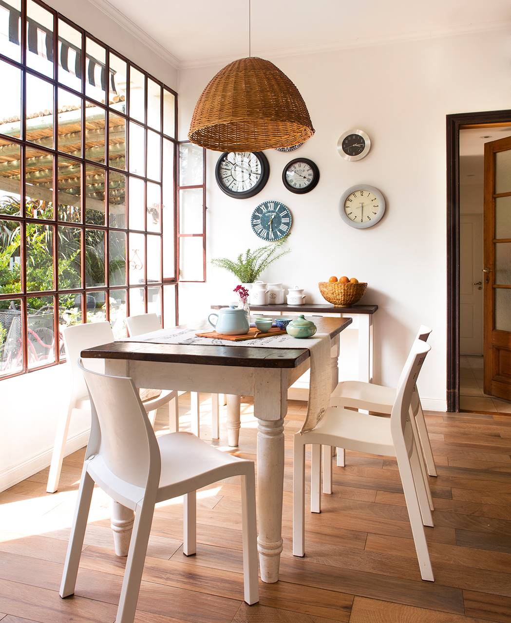 Comedor con mesa blanca de madera, ventana y relojes en la pared.