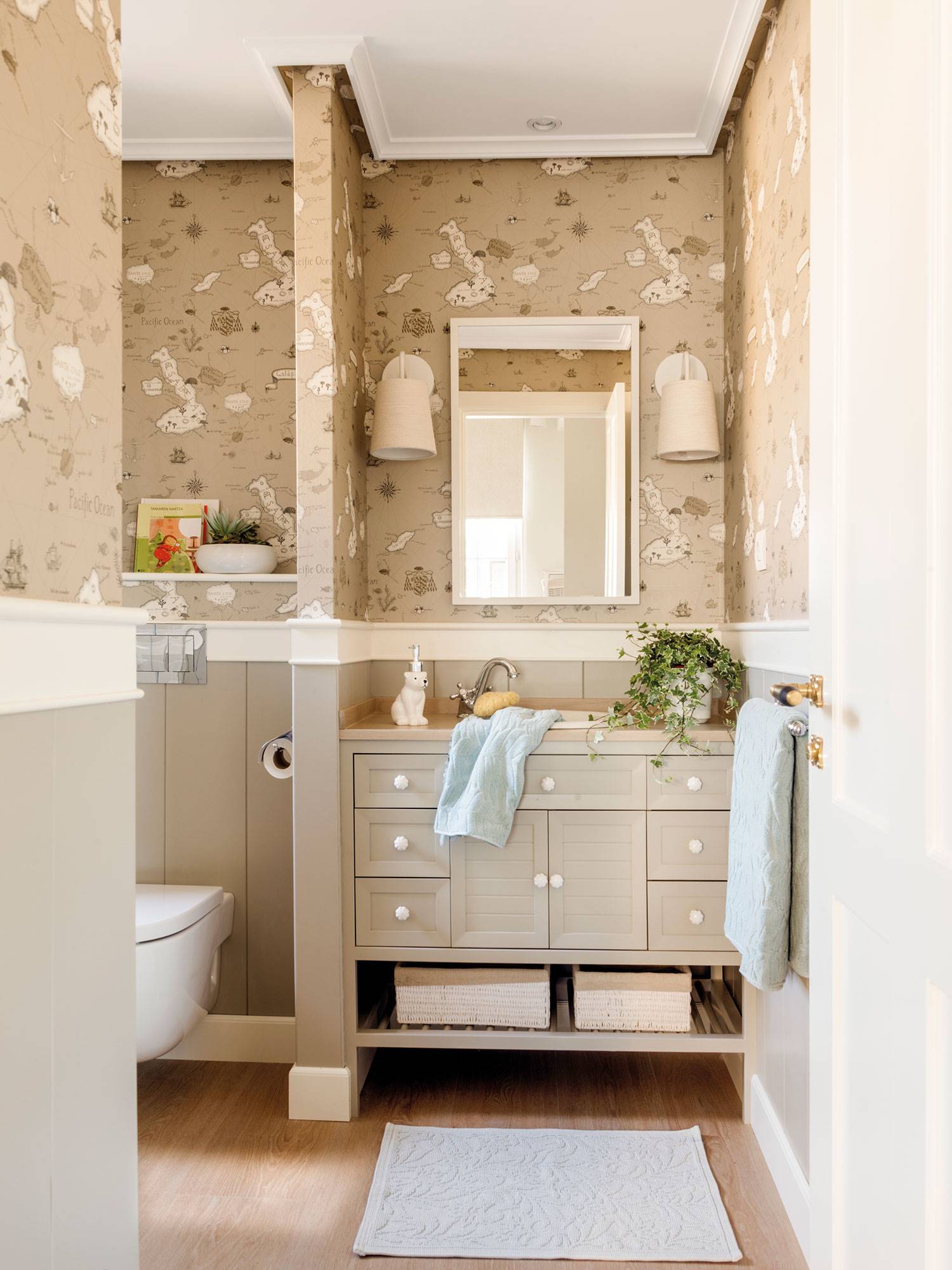 Baño con mueble de color beige, papel pintado y murete que separa la zona del inodoro. 