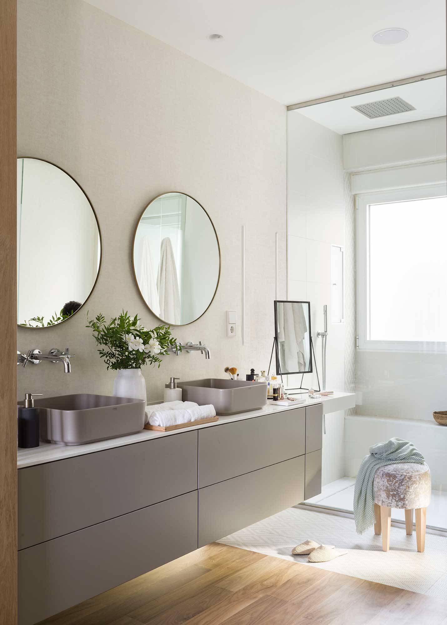 Baño con mueble gris con encimera blanca y dos lavamanos grises y espejos redondos. 