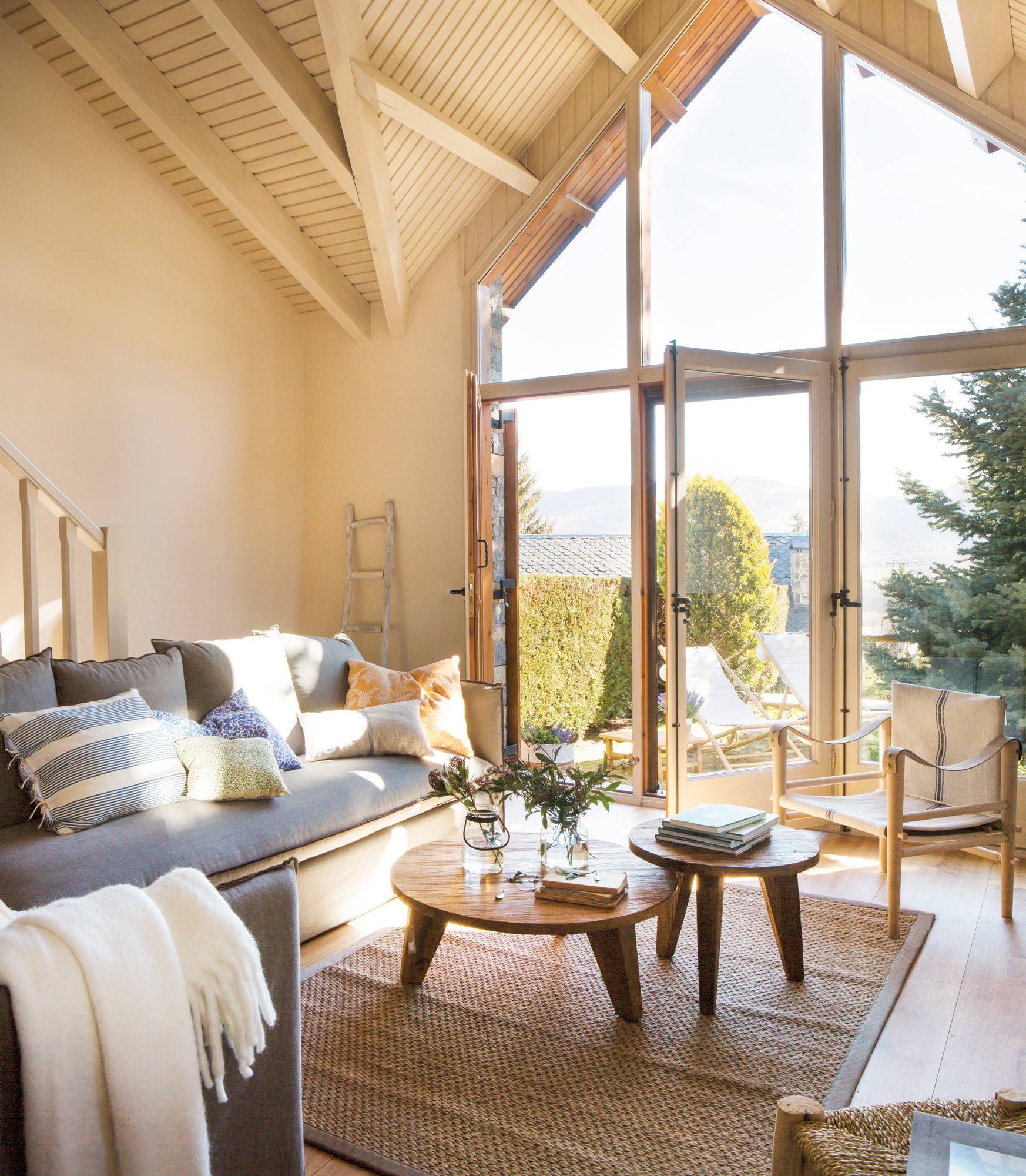 Salón pequeño de casa de montaña con sofá gris, mesas de centro de madera y gran ventanal hasta el techo.