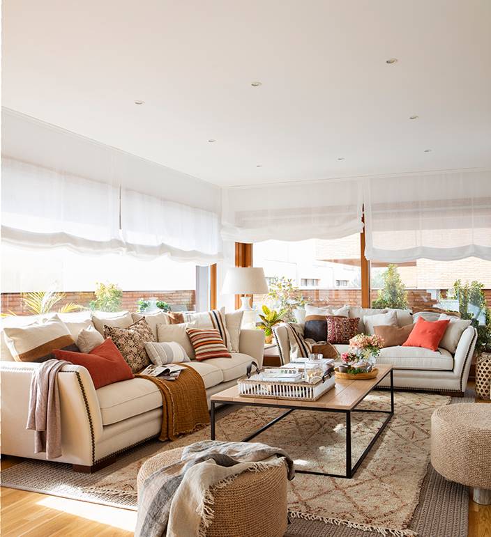 Salón clásico con sofás beige con tachuelas, mesa de centro de madera y estores blancos en las ventanas.