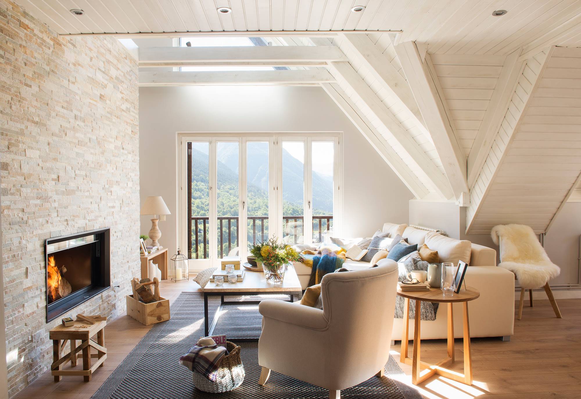 Salón de casa de montaña con paredes y techos de madera pintada de blanco, pared de piedra con chimenea y sofá beige. 