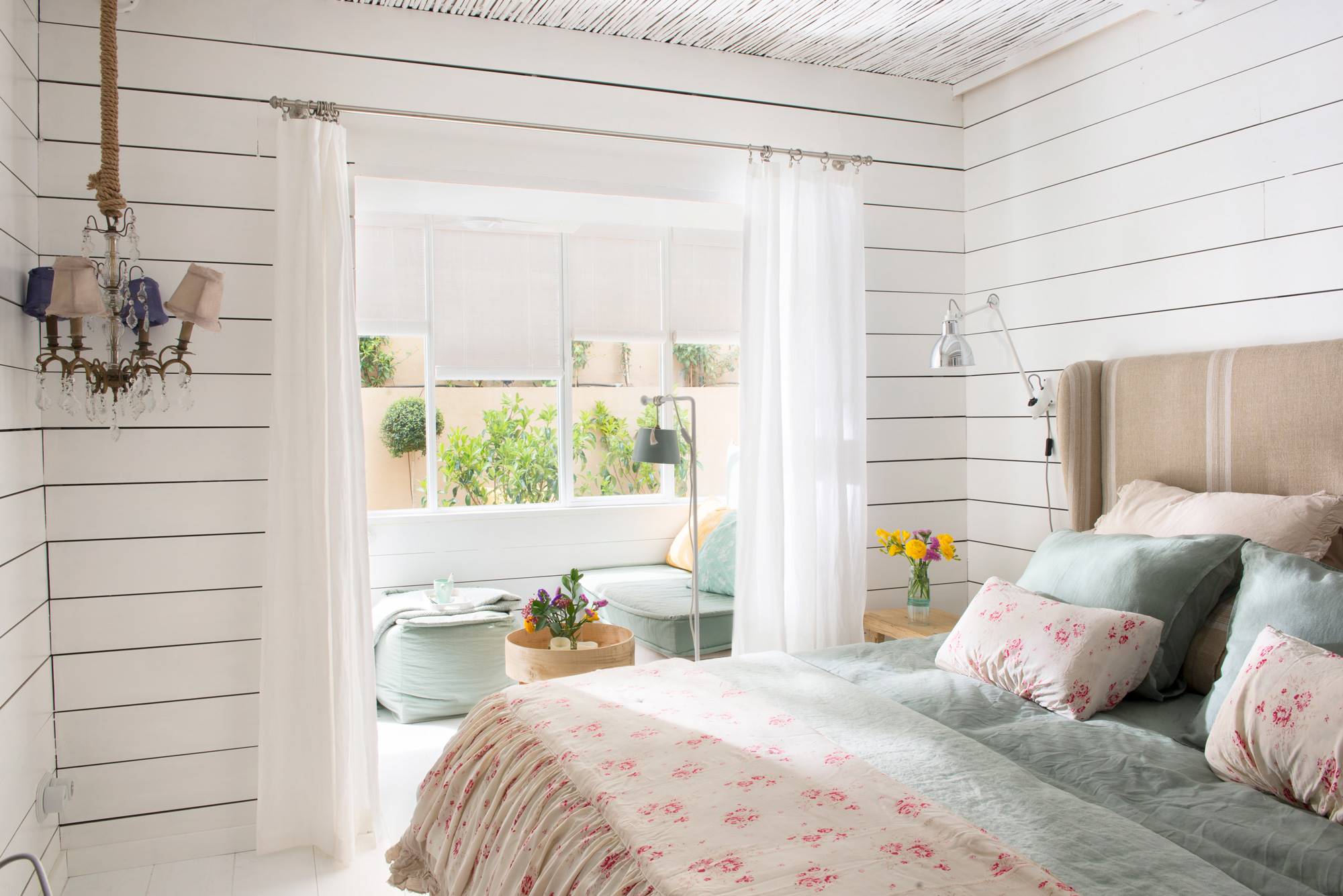 Dormitorio de primavera con paredes de lamas horizontales en blanco, sillón bajo la ventana y ropa de cama de flores