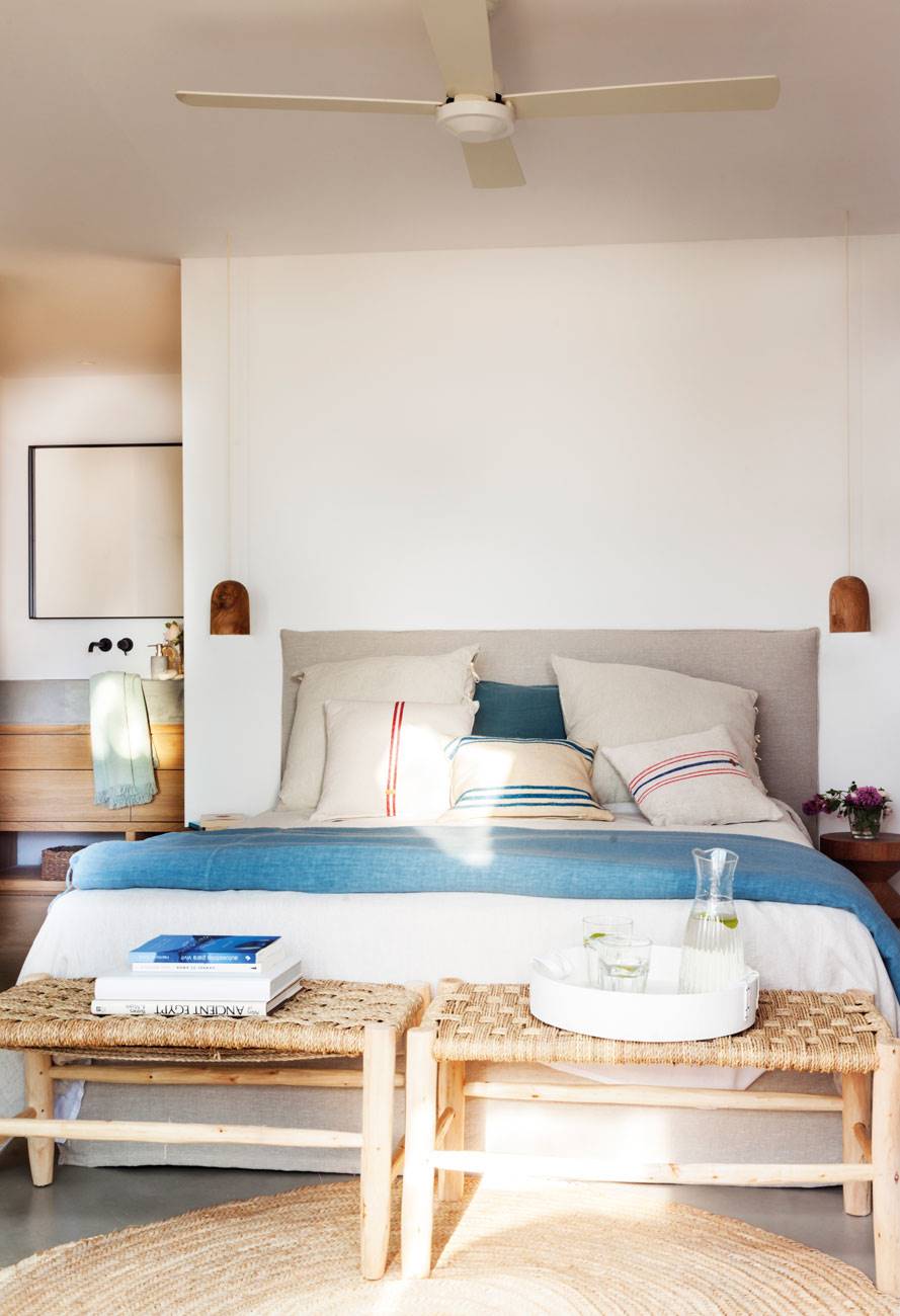 Dormitorio de primavera decorado en blanco y azul con bancos de enea y madera a los pies.