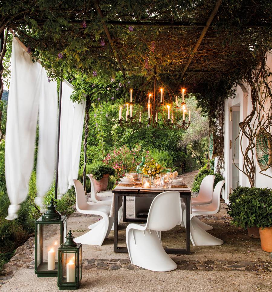 Comedor exterior elegante con cortinas, iluminación y plantas enredaderas encima de la pérgola.