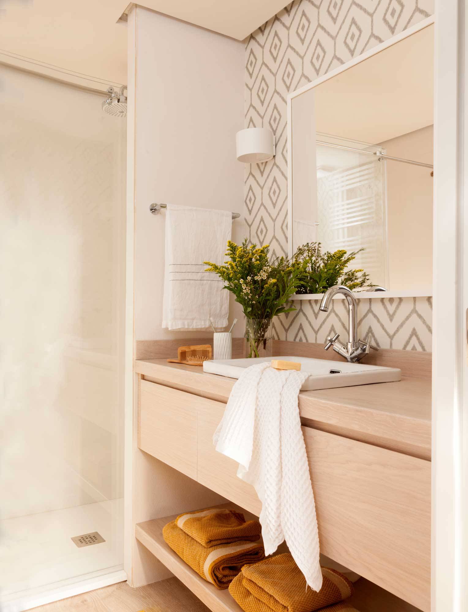 Baño con mueble bajolavabo de madera, espejo, pared con papel pintado geométrico y ducha. 