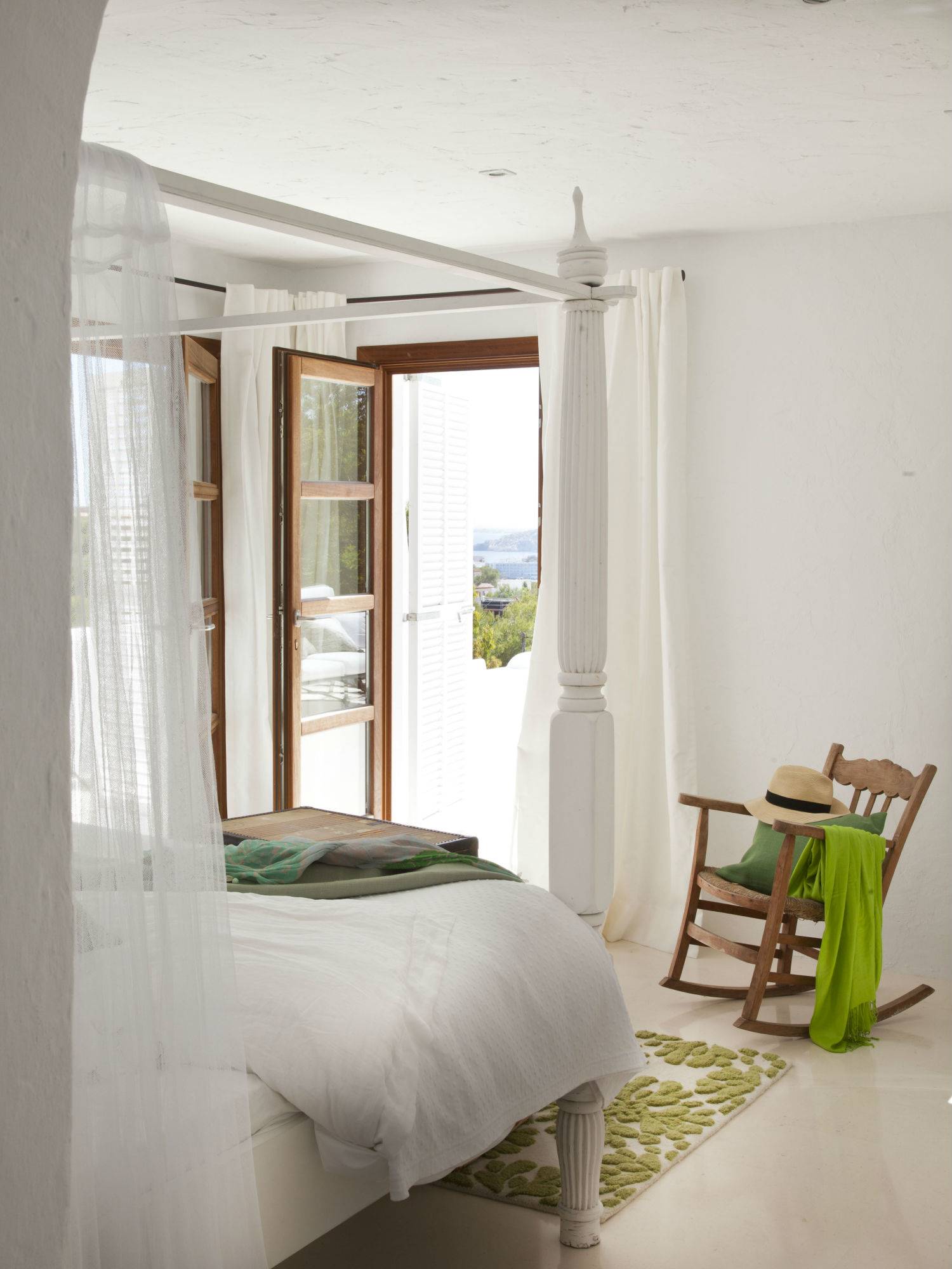 Dormitorio veraniego con cama lacada en blanco, silla mecedora en madera y con vistas al mar.