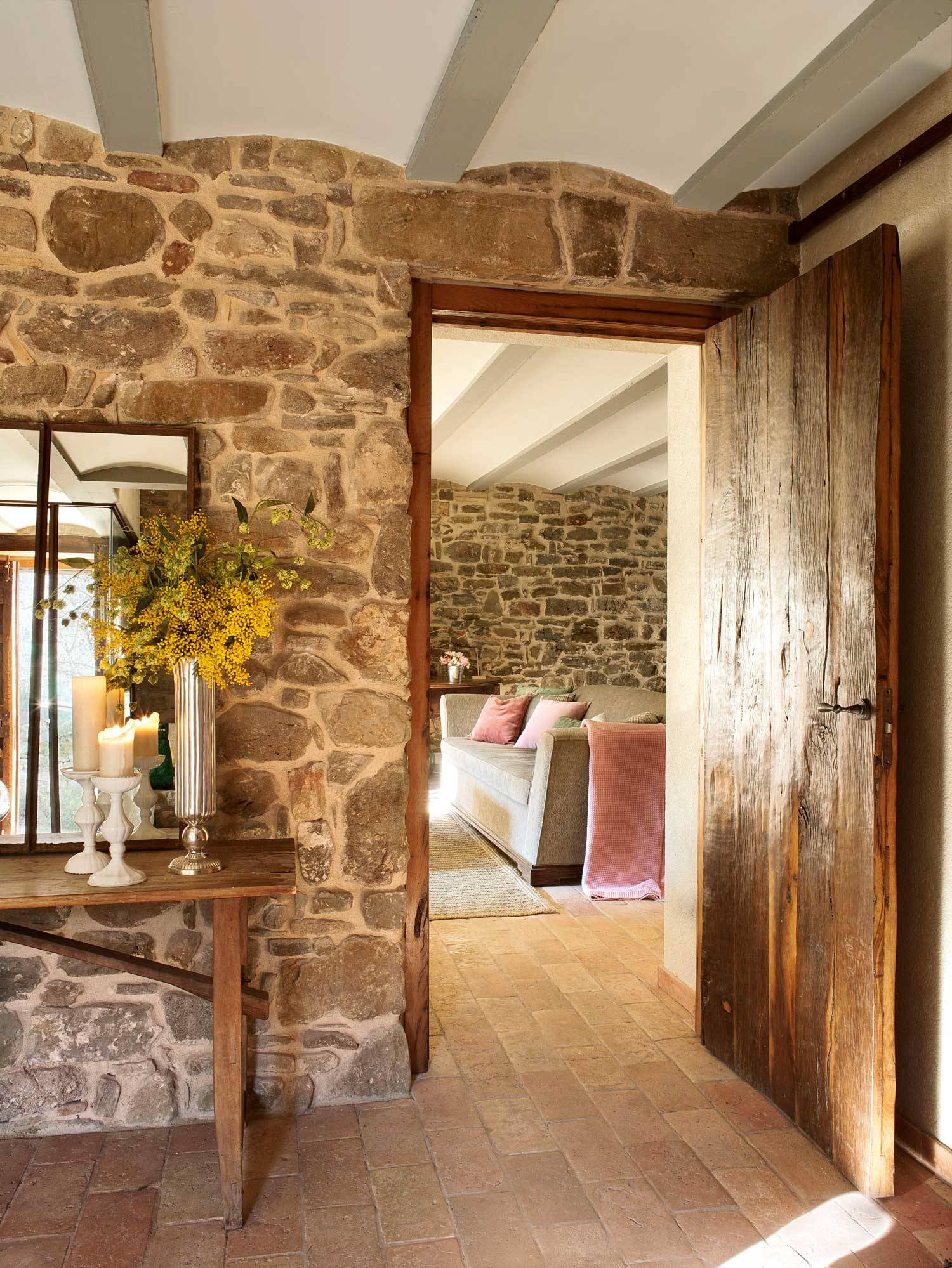 Recibidor rústico con pared de piedra, puerta de madera, consola de madera, espejo, velas y mimosas-360215