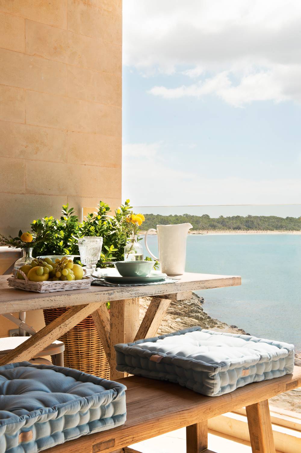 Comedor exterior en la terraza con vistas al mar, mesa y banco de madera y cojines de asiento de color azul.