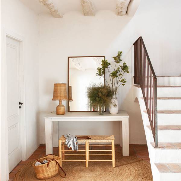 Recibidor rústico con suelo de toba, vigas blanqueadas, escalera, consola, esoejo, banco, cesta y alfombra de fibra y pared blanca-510271