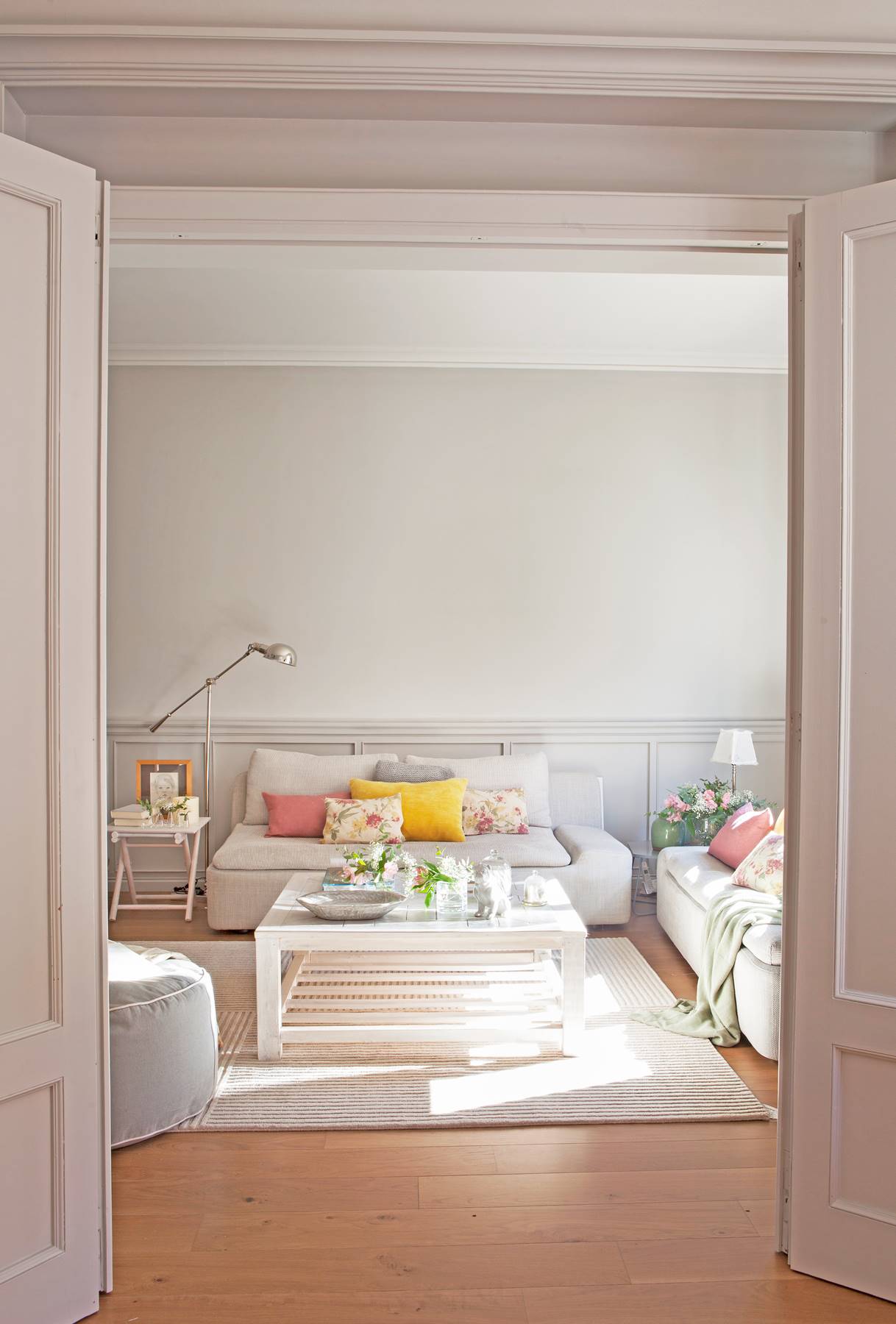 Salón con pared y sofá gris, suelo de parquet, mesa de centro de madera blanca, cojines amarillos y rosas y estampados de flores_423868