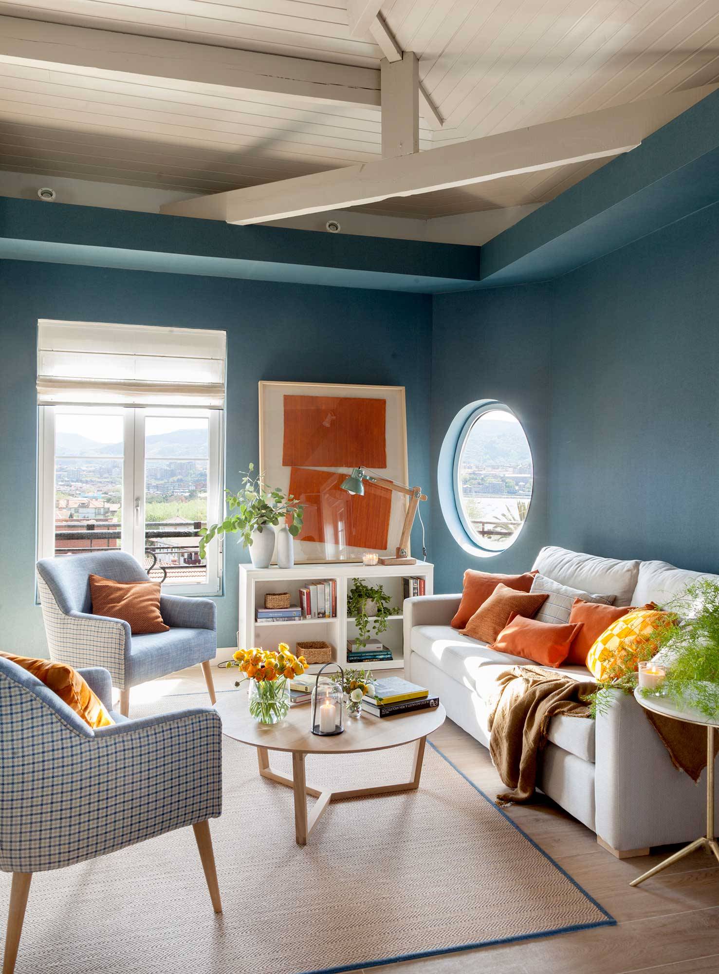 Salón pequeño con paredes azules, vigas en el techo, butacas y mesa de centro de madera. 