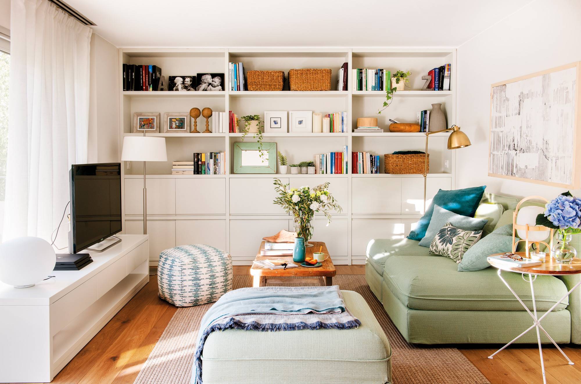 Salón pequeño con librería blanca de pared a pared y sofá verde con puff a juego. 