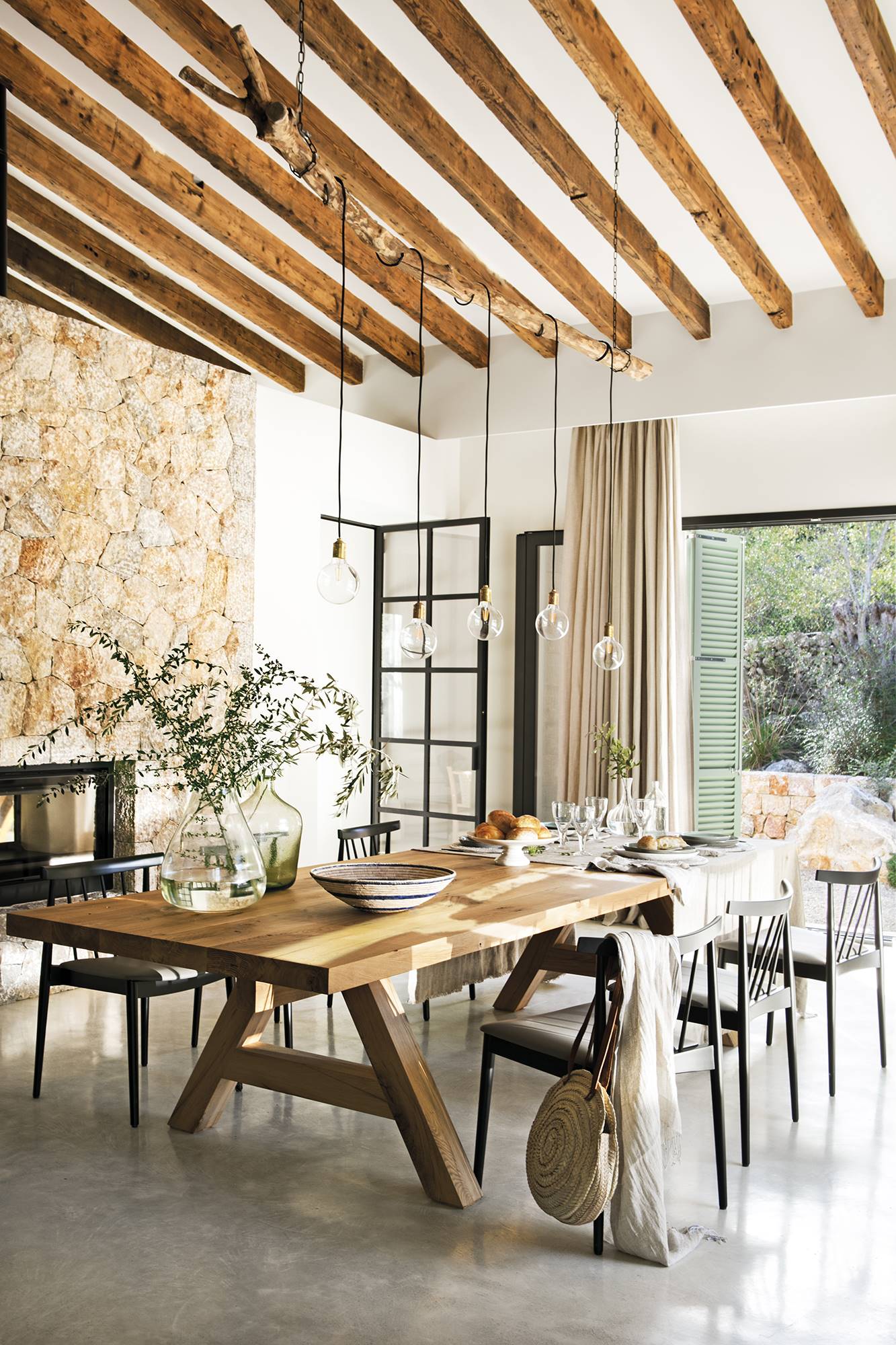 Comedor con aire rústico y de campo con mesa de madera, sillas, pared de piedra y vigas en el techo.