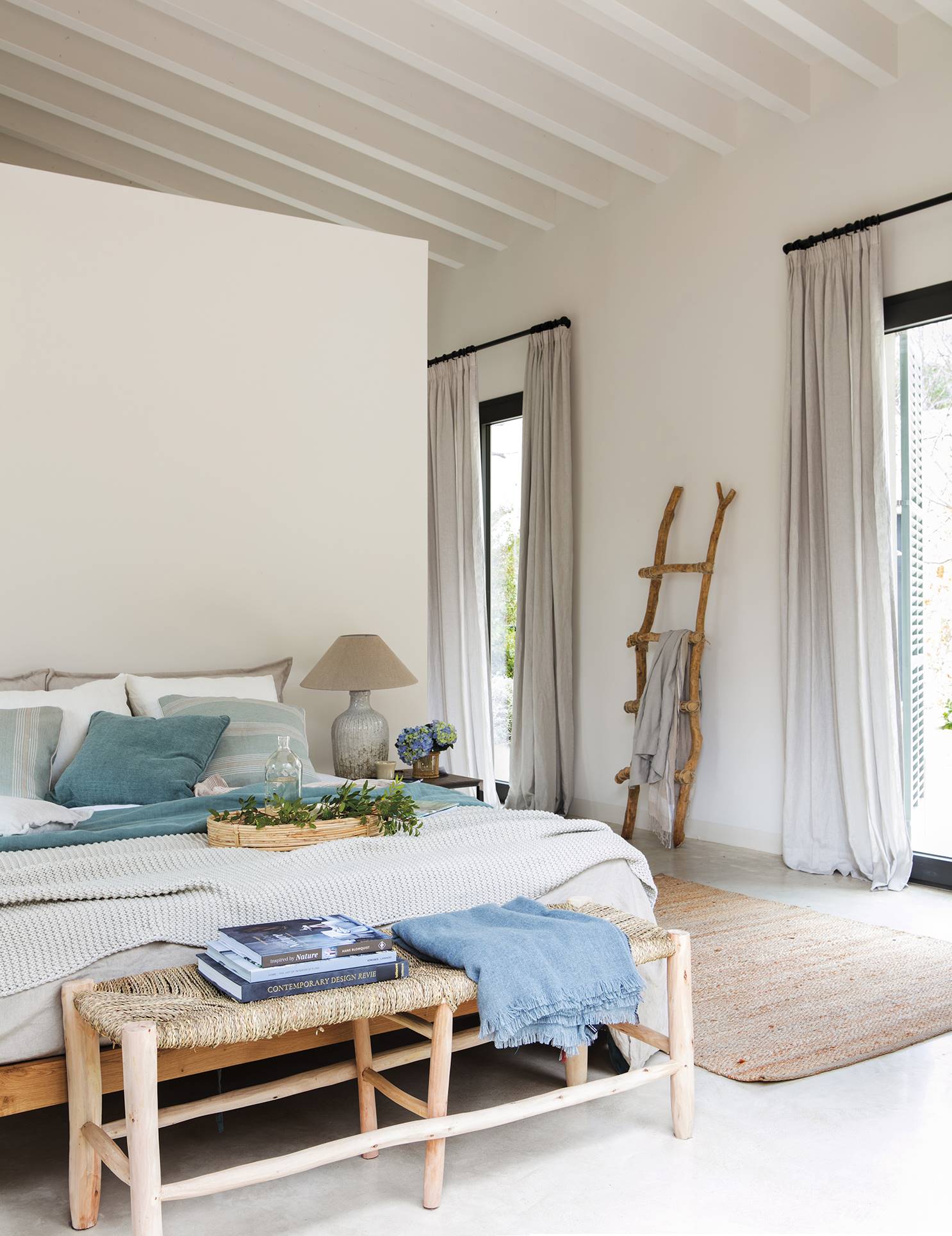 Dormitorio veraniego con textiles ligeros en la cama, banqueta al pie de la cama de fibras y escalera al fondo decorativa.