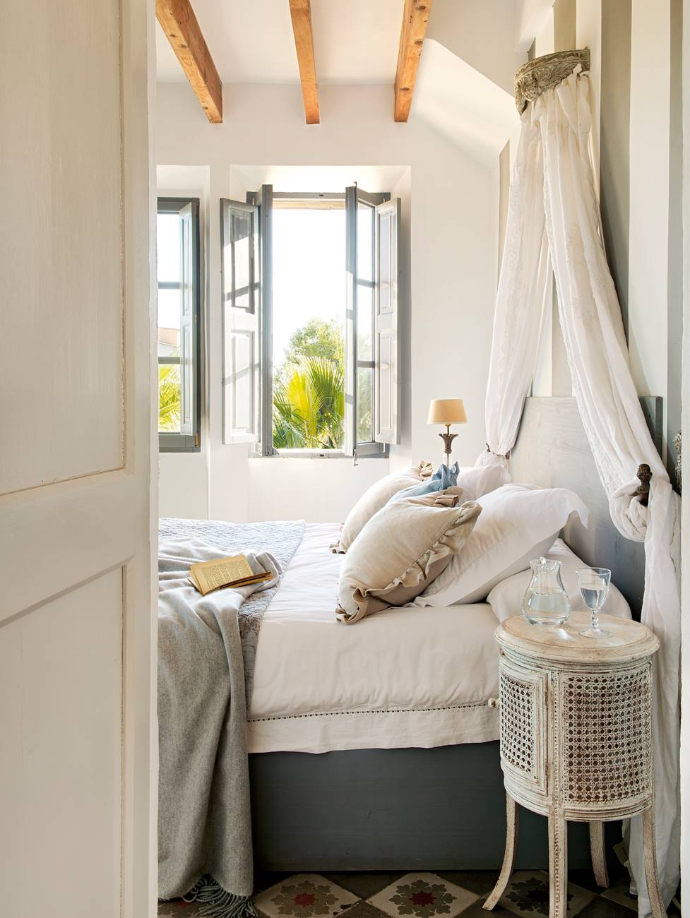 Dormitorio con mesilla de noche de mimbre, vigas de madera, ventana con porticones de color azul y mosquitera
