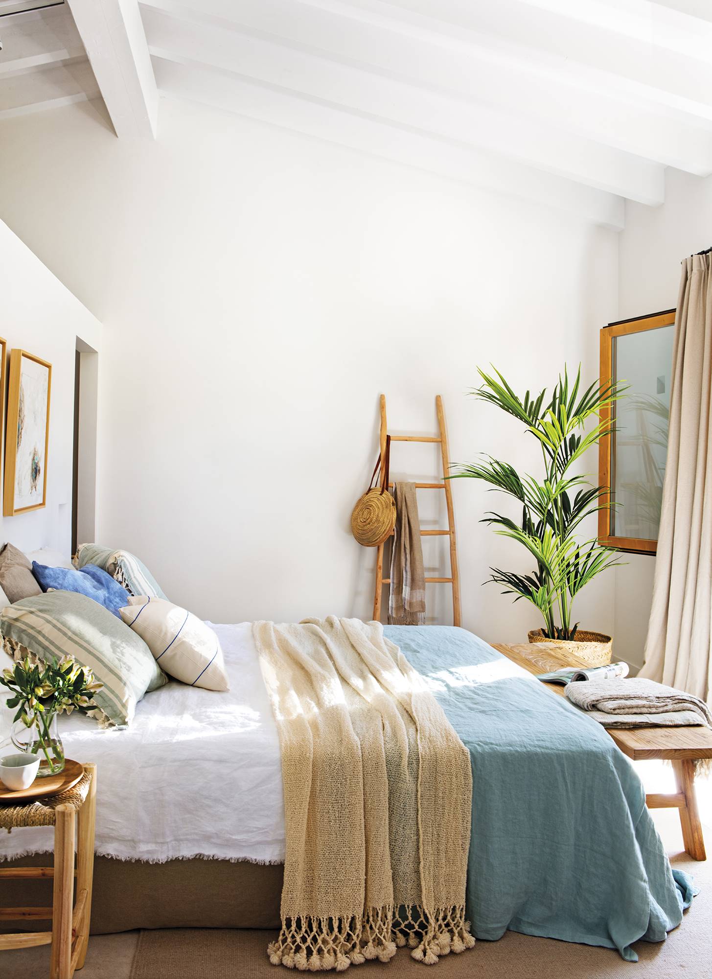 Dormitorio con textiles de cama en beige y azul, taburete como mesilla de noche, banco de madera a los pies y escalera decorativa